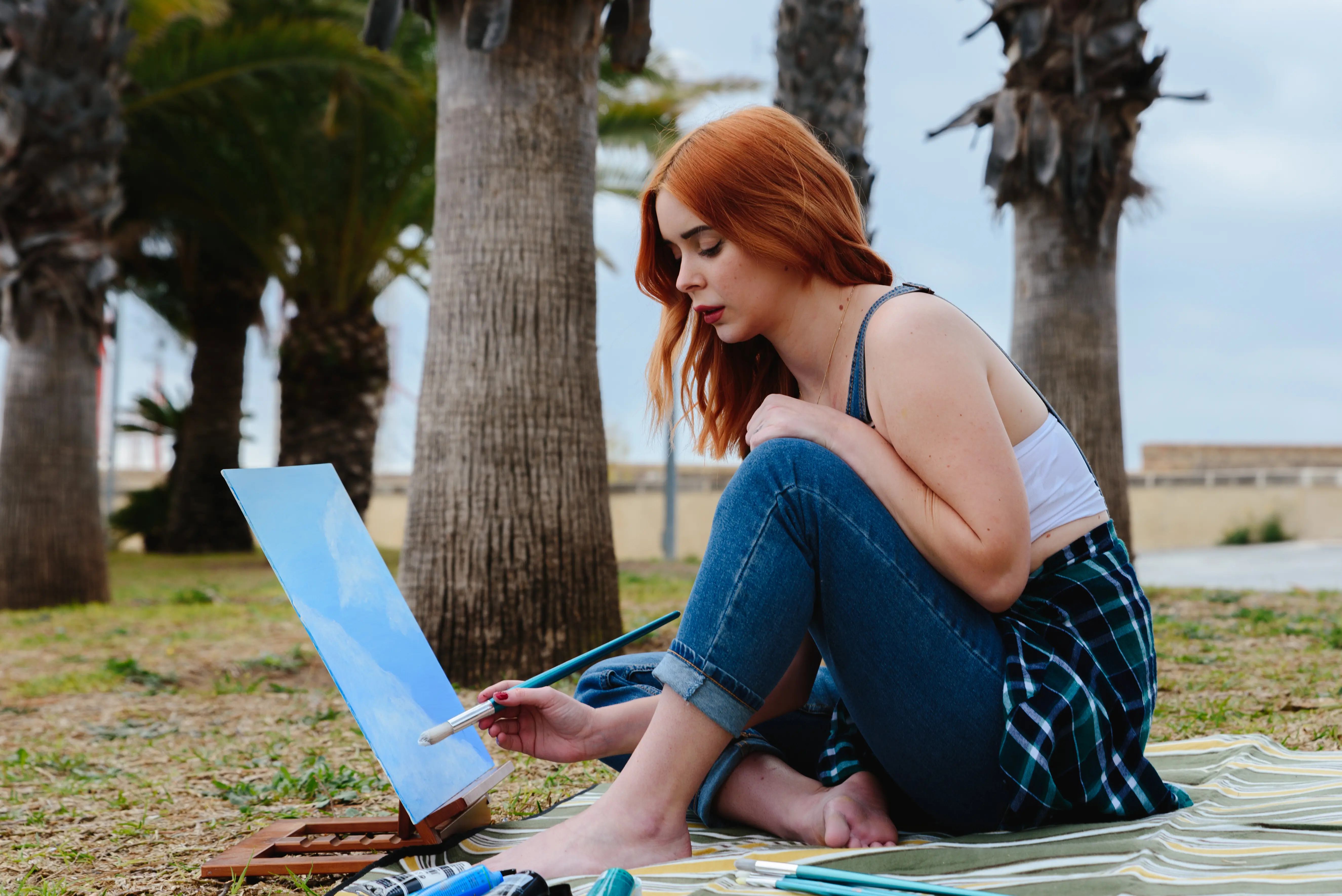 Eine junge kaukasische rothaarige Künstlerin malt draußen im Park mit einem Pinsel an einer Leinwand auf einer kleinen Staffelei.