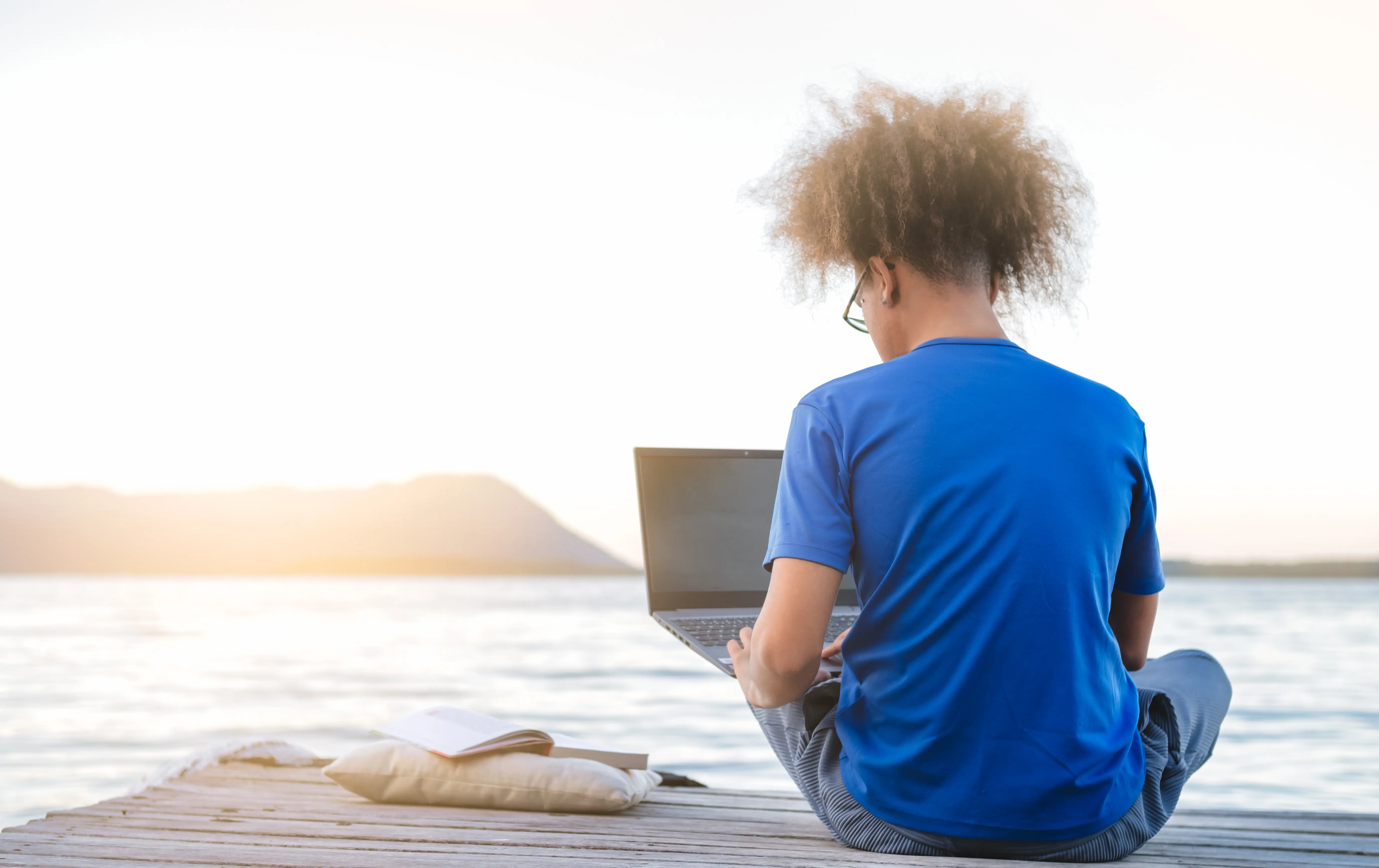 Junger hipsterhafter Mann, ein Digitalnomade, sitzt auf einem Holzsteg am Meer und arbeitet bei Sonnenuntergang aus der Ferne im Internet; Reisen mit einem Computer; Konzept eines Online-Traumjobs; selektiver Fokus.